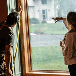 A contractor measures a window while talking with a homeowner.