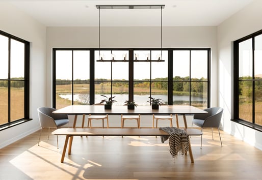 A simply furnished dining space with black windows on three sides that afford views of a prairie and wetland.
